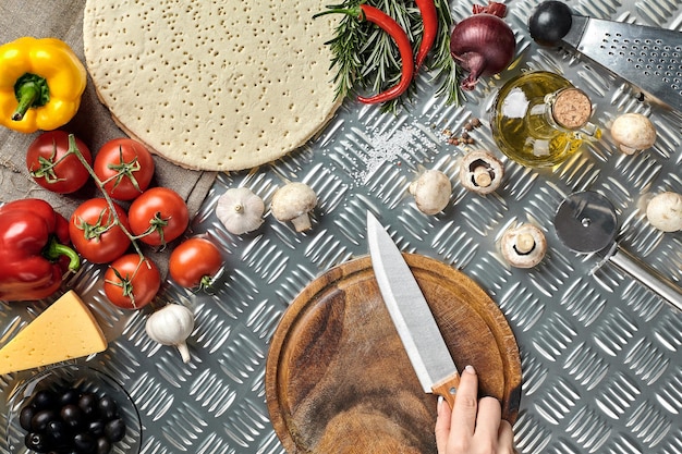 Mano femminile con coltello sul tavolo da cucina in metallo intorno giacciono ingredienti verdure e spezie per pizza