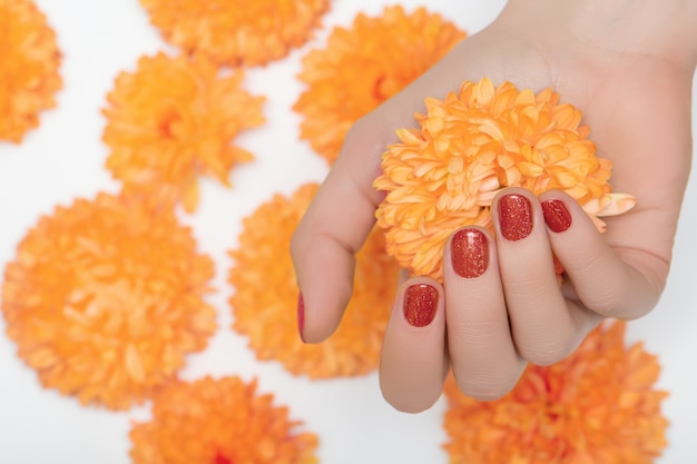 Mano femminile con il design delle unghie rosso glitter che tiene il fiore dell'orchidea arancione.