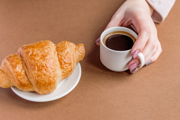 Mano femminile con il manicure che tiene tazza di caffè e che mangia croissant. Colazione in stile francese