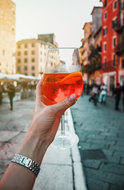 Mano femminile con un bicchiere di spritz da cocktail arancione vicino a vecchi edifici Giorno soleggiato a Verona, in Italia