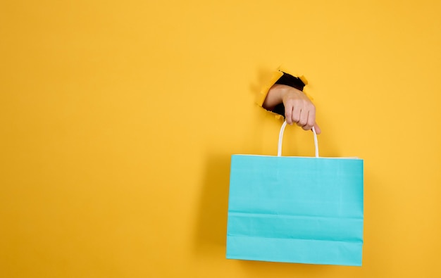 Mano femminile con una borsa della spesa di carta blu che spunta da un foro strappato su uno sfondo di carta gialla. Rifiuto della plastica, inizio delle vendite