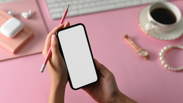Mano femminile creativa che tiene il mockup dello schermo bianco dello smartphone e la matita sulla scrivania rosa dell'ufficio