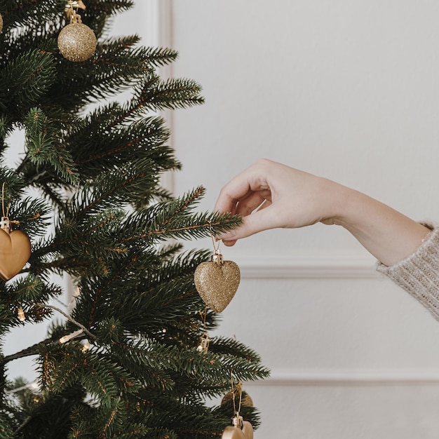 Mano femminile decorare bellissimo albero di Natale con palline dorate e giocattoli davanti al muro bianco Elegante sfondo estetico Natale Capodanno vacanze celebrazione preparazione