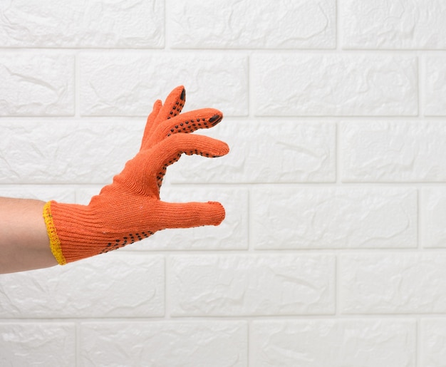 mano femminile in un guanto protettivo da lavoro arancione sullo sfondo di un muro di mattoni bianchi, la mano tiene condizionatamente un oggetto