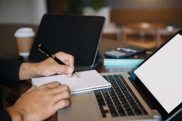 Mano maschile che prende appunti su un taccuino scritto a mano che scrive un programma di lavoro e usa un computer portatile alla scrivania dell'ufficio al mattino