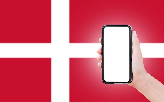 Mano maschio che tiene smartphone con spazio vuoto sullo schermo sullo sfondo della bandiera sfocata della Danimarca Primo piano