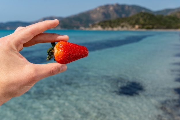 Mano ravvicinata che tiene tra le dita una fragola matura con una bella acqua di mare traslucida idilliaca.