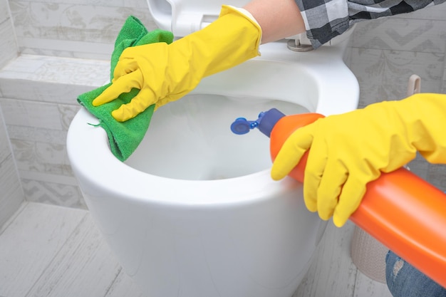 Mano ravvicinata con detergente per la pulizia della toilette Servizio di pulizia mani ravvicinate che indossano guanti protettivi gialli utilizzando una soluzione detergente liquida per la pulizia della toilette con sciacquone, disinfezione e concetto di igiene