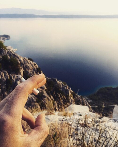 Mano tagliata di una persona che tiene una sigaretta sul mare contro il cielo