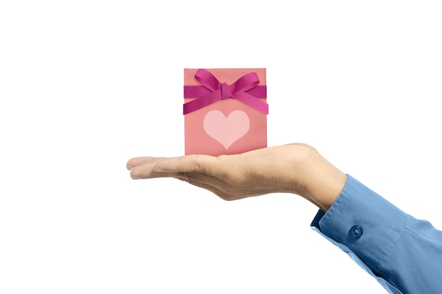 Mano umana che mostra confezione regalo rosa con nastro rosso e cuore isolato sopra il muro bianco