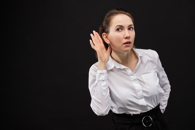 Mano vicino all'orecchio, stagno per sentire qualcosa. Ragazza in una camicia bianca su sfondo nero.