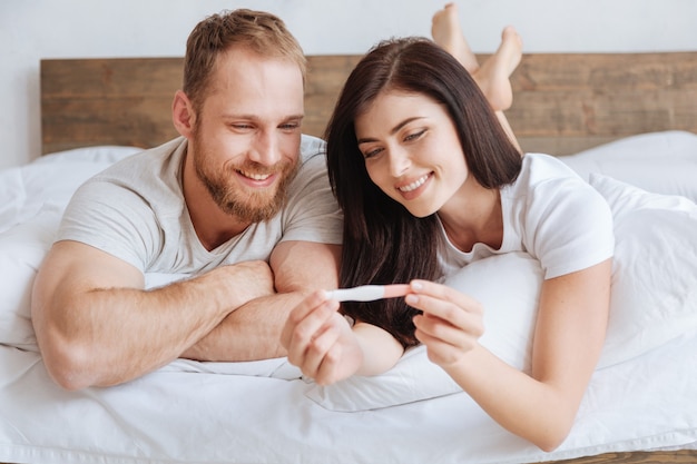 Marito e moglie appena sposati sorridono ampiamente mentre guardano un test di gravidanza positivo a letto
