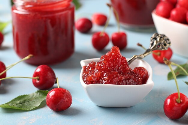 Marmellata fatta in casa di ciliegie in una ciotola bianca e vasetti su sfondo azzurro. Avvicinamento