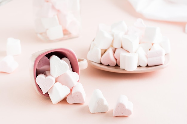 Marshmallow bianchi e rosa a forma di cuore sono sparsi da una tazza e su un piattino su uno sfondo rosa