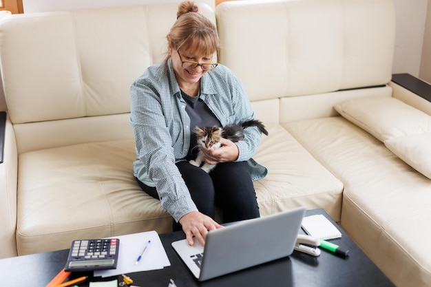 Marure donna in camicia blu seduta con un gatto in grembo al tavolo di legno a casa con laptop e notebook che lavorano o fanno acquisti online