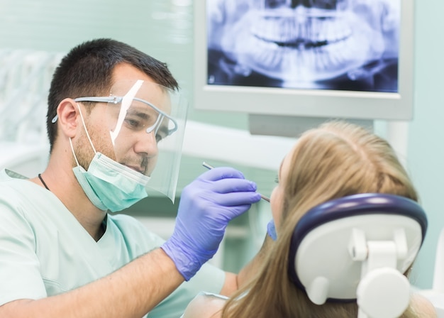 Maschera del primo piano della giovane donna che si siede nella sedia del dentista con la bocca aperta all'ufficio del dentista mentre avendo esame.