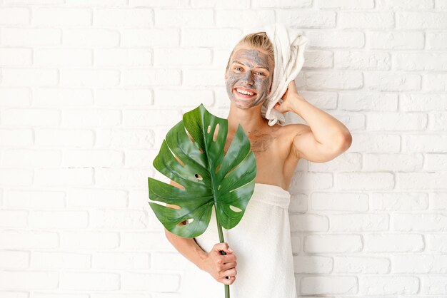 Maschera facciale spa. Spa e bellezza. Donna sorridente felice che indossa asciugamani da bagno con una maschera facciale di argilla sul viso che tiene una foglia di monstera verde