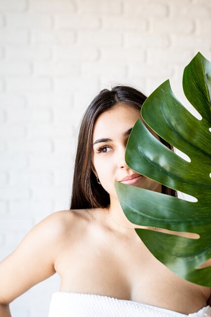 Maschera facciale spa. Spa e bellezza. Felice bella donna bruna indossando asciugamani da bagno tenendo una foglia di monstera verde davanti al viso