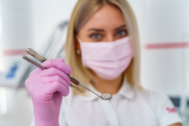 Maschera per camice rosa sul viso di una dottoressa. Strumenti per l'esame della cavità orale nelle mani.