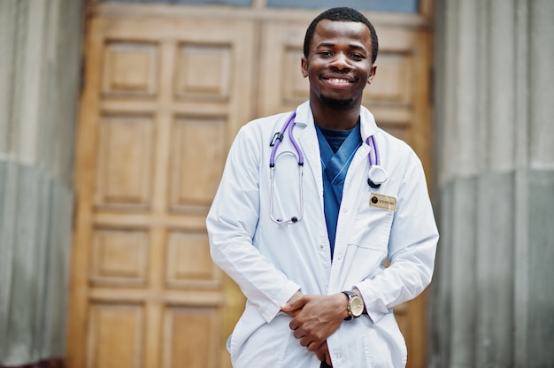 Maschio afroamericano di medico al cappotto del laboratorio con lo stetoscopio all'aperto contro la porta della clinica.