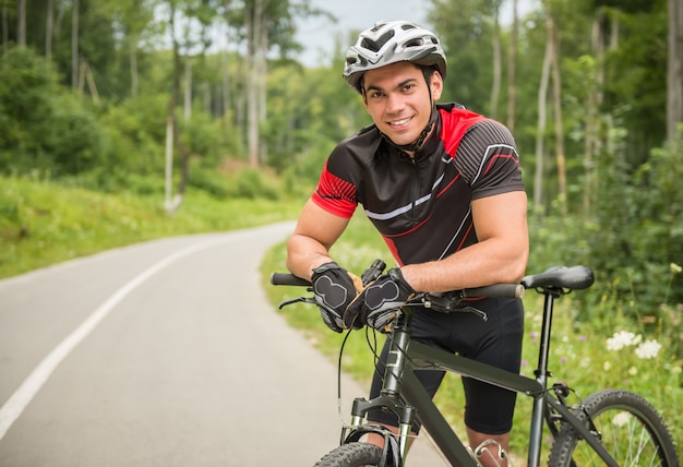 Maschio allegro del ciclista che si appoggia la sua bici sul sentiero forestale