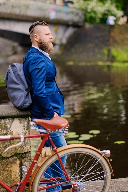 Maschio barbuto rosso vestito con una giacca blu e jeans su una bicicletta retrò in un parco.