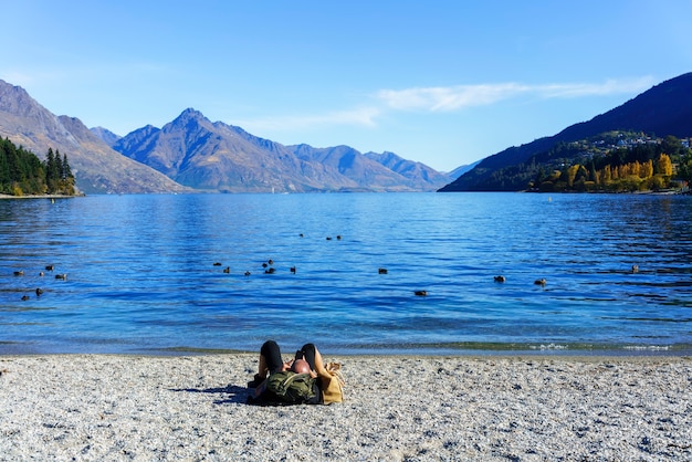 Maschio caucasico utilizzando smart phone e rilassarsi sulla spiaggia del lago Wakatipu, Queenstown, Isola del Sud della Nuova Zelanda