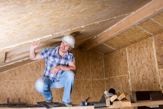 Maschio lavoratore edile Builder bianco da indossare elmetto accovacciato su ponteggi sopraelevati vicino al soffitto in casa incompiuta con pannelli di compensato di particelle esposte