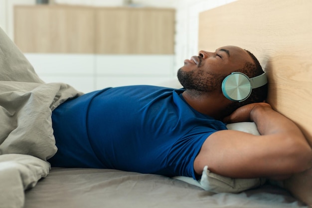 Maschio nero sdraiato indossando le cuffie ascoltando musica rilassante in camera da letto