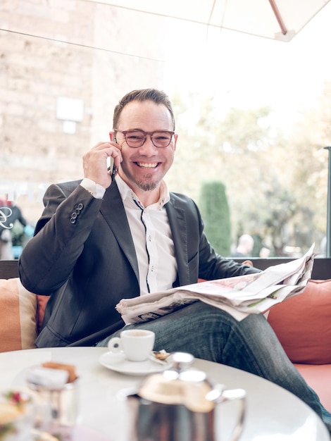 Maschio sorridente con giacca nera e occhiali seduto in un caffè con un giornale e una tazza di caffè mentre parla al cellulare e guarda la telecamera