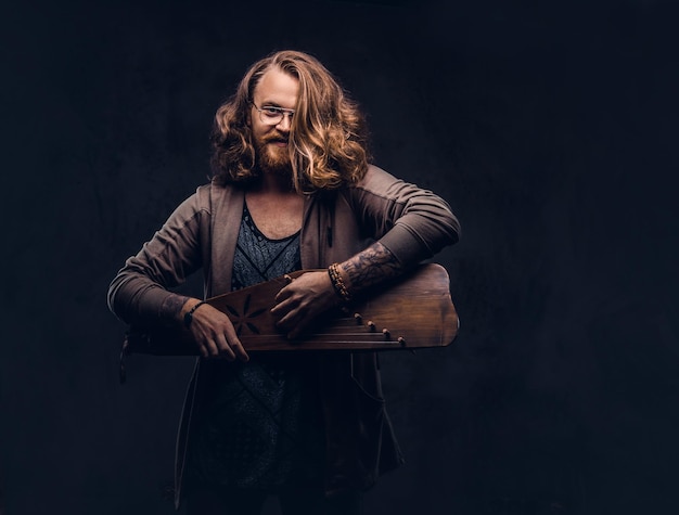 Maschio sorridente dei pantaloni a vita bassa della testarossa con i capelli lunghi e lussureggianti e la barba piena vestito in abiti casual che giocano su uno strumento musicale tradizionale russo - gusli. Isolato su sfondo scuro.