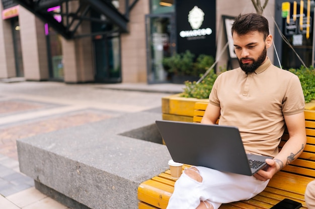 Maschio studente barbuto concentrato che studia digitando con il computer portatile seduto su una panchina sulla strada della città con caffè da asporto