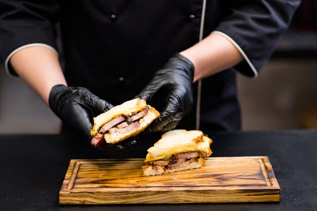 Masterclass di cucina. Primo piano delle mani dello chef che tengono una metà del sandwich di petto di manzo affumicato con formaggio su tavola di legno.