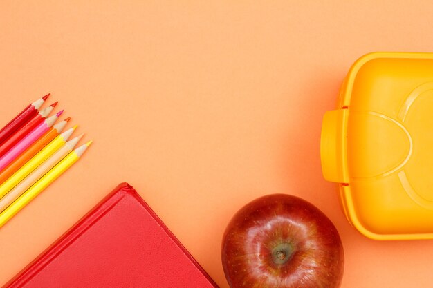 Materiale scolastico. Matite colorate, libro, mela e scatola per il pranzo su sfondo rosa. Vista dall'alto con copia spazio. Torna al concetto di scuola. Colori pastello