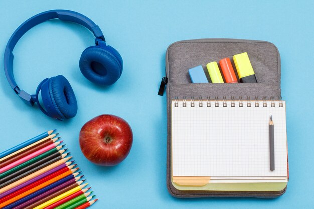 Matite colorate, mela, cuffie, quaderno aperto su astuccio portamatite con pennarelli colorati e pennarello su sfondo blu. Vista dall'alto. Torna al concetto di scuola. Materiale scolastico.