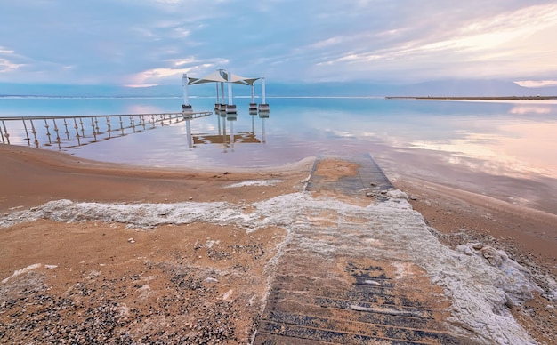 Mattina calma sulla spiaggia del Mar Morto di Ein Bokek, nuvole rosa riflesse sulla superficie dell'acqua vicino a binari in acciaio che conducono al riparo del sole, sabbia ricoperta di cristalli di sale