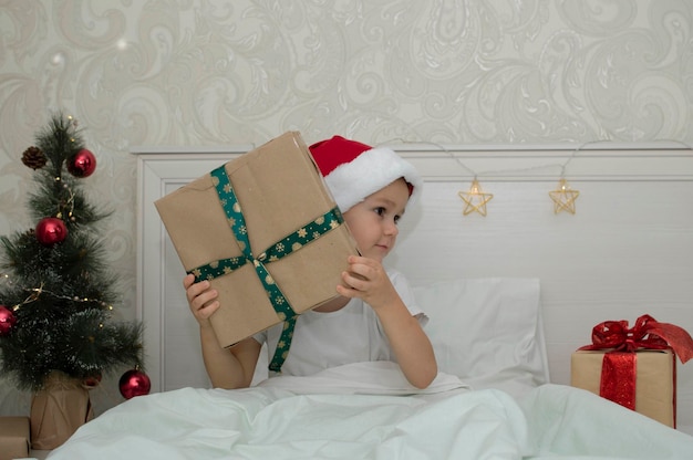 Mattina di Natale, bambina in pigiama con regali a letto sullo sfondo dell'albero di Natale. Un bambino felice e sorridente apre il regalo di Capodanno a casa. Il concetto di vacanze, Capodanno, Natale