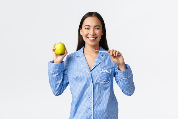 Mattina, stile di vita attivo e sano e concetto di casa. Sorridente bella ragazza asiatica che mostra spazzolino da denti e mela verde, ha denti bianchi perfetti e dà consigli sull'igiene della bocca, sfondo bianco