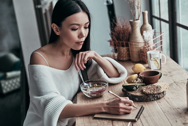 mattinata impegnativa. attraente giovane donna che mangia una sana colazione e scrive qualcosa