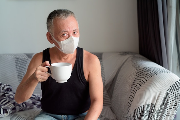 Maturi l'uomo giapponese con la maschera che si siede e che beve il caffè a casa sotto la quarantena