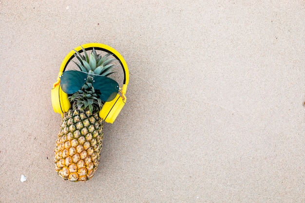 Maturo attraente ananas in eleganti occhiali a specchio e cuffie d'oro sulla sabbia contro l'acqua di mare turchese. Concetto di vacanze estive tropicali.
