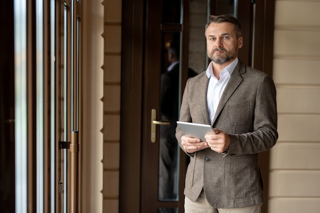 Maturo imprenditore barbuto con touchpad ti guarda mentre sei in piedi e networking all'interno del business center
