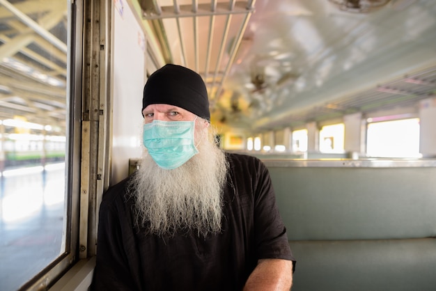 Maturo uomo turistico barbuto con maschera per la protezione dall'epidemia di virus corona seduto all'interno del treno