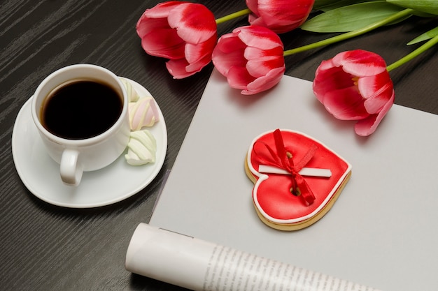 Mazzo di tulipani rosa, una tazza di caffè, biscotti a forma di cuore rossi con una nota, rivista vuota