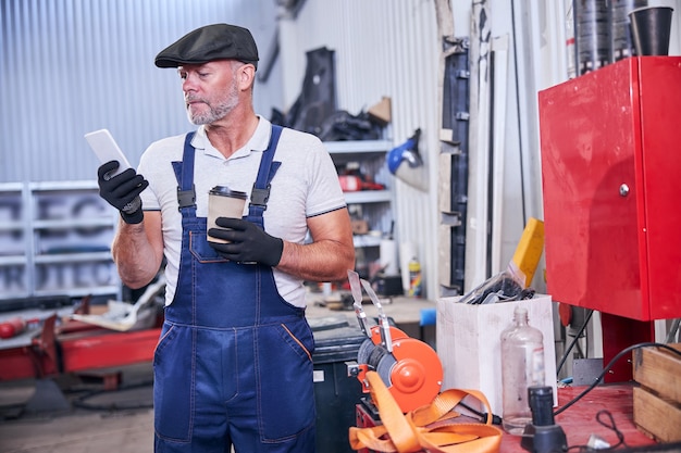 Meccanico barbuto che utilizza il telefono cellulare nell'officina di riparazioni auto