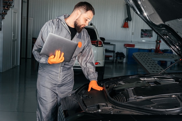 Meccanico di automobile che lavora con un computer portatile nel servizio di riparazione automatica che controlla il motore dell'automobile