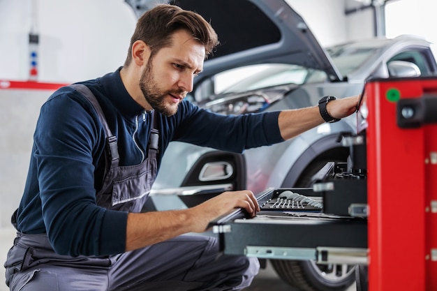 Meccanico esperto dedicato che si accovaccia accanto alla scatola con gli strumenti e sceglie lo strumento giusto per riparare l'auto. Garage interno del salone dell'auto.