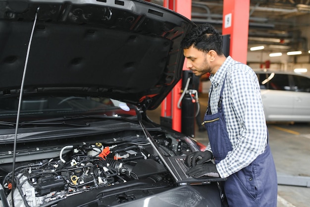 Meccanico uomo manager meccanico lavoratore utilizzando un computer portatile controllo auto in officina presso il centro di servizio di riparazione auto ingegnere giovane guardando i dettagli del veicolo di ispezione sotto il cofano dell'auto