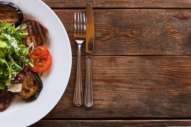 Medaglioni con verdure grigliate su piatto bianco Carne pomodori melanzane e insalata verde con formaggio grattugiato che serve nel ristorante vista dall'alto con spazio libero su sfondo di legno a destra