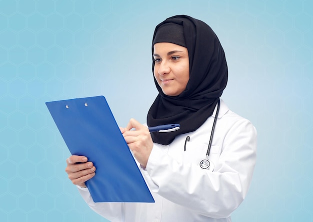 medicina, assistenza sanitaria e concetto di persone - sorridente medico musulmano donna che indossa hijab e cappotto bianco con clipboard e stetoscopio su sfondo blu